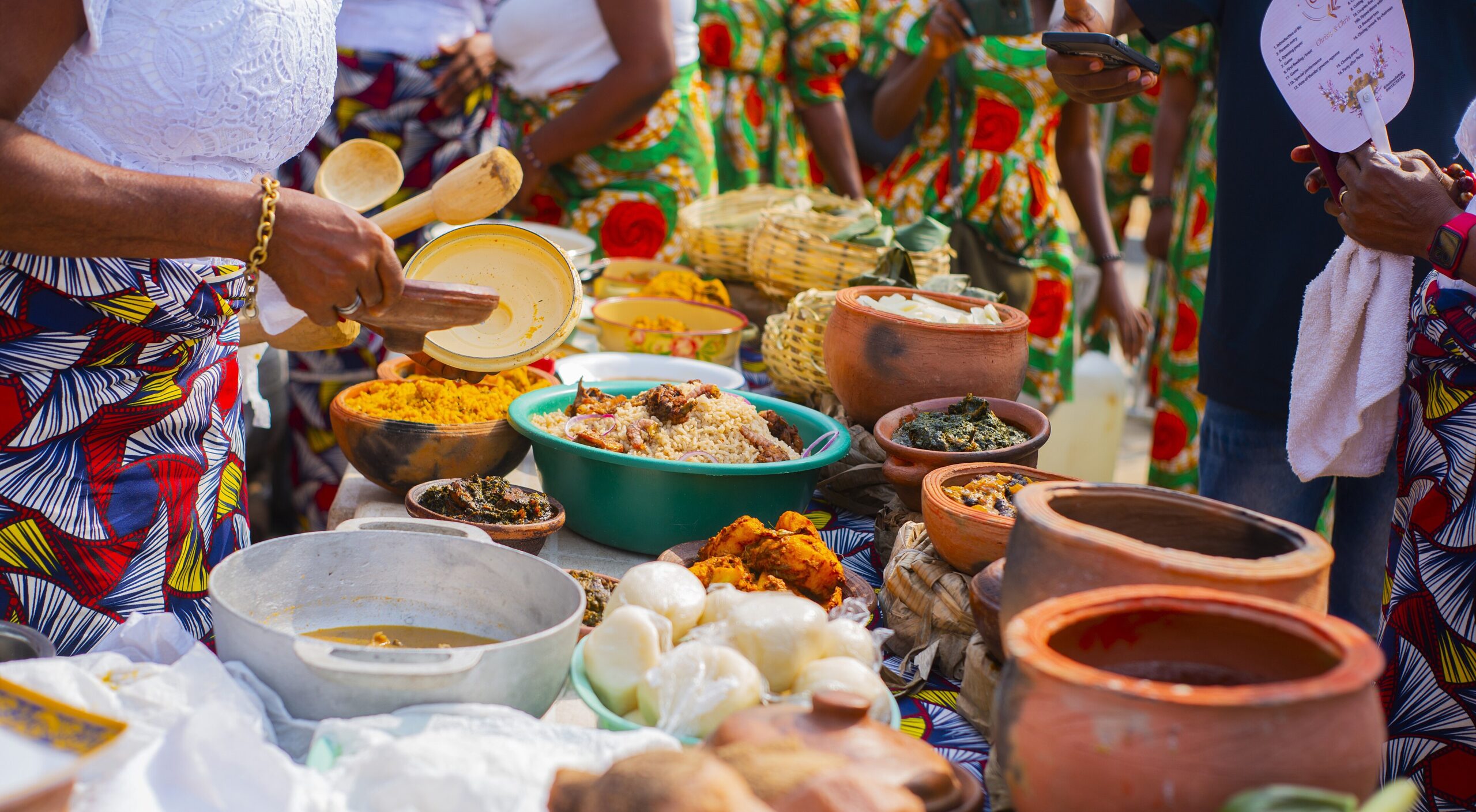 A table of traditional Nigerian dishes in clay pots and bowls, with women in Ankara serving food