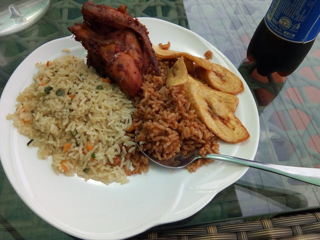 Plate of jollof rice and fried rice with fried plantain and chicken, with a bottled drink